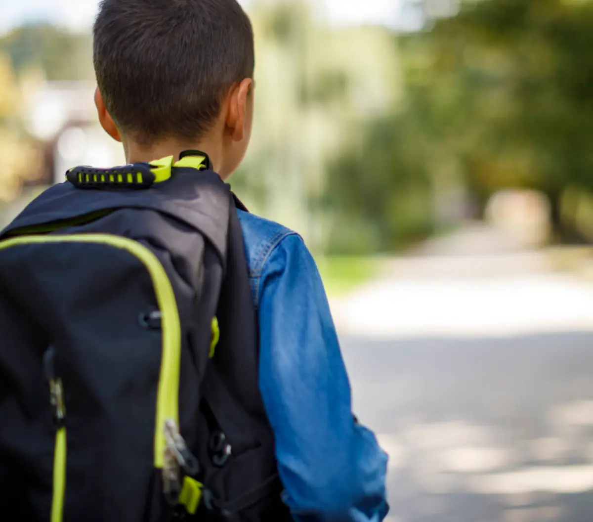 Cropped digital stock photograph image of a young boy seen from behind, wearing a black backpack with lime green accents and a blue denim shirt as he walks and glances down a concrete path that is surrounded by trees in the blurred background around him