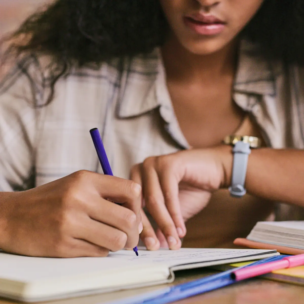 Cropped digital stock photograph image of a female individual with long curly dark black hair and a beige plaid shirt is focusing on writing in an open notebook with a purple pen; A watch is on her left wrist, and other various random assorted pens and a book are on the table beside her