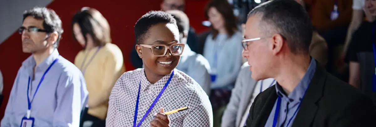 Cropped digital stock photograph image of a smiling Black woman with short black hair and big beige colored outer frame prescription eyeglasses wearing a patterned button-up shirt and a blue lanyard as she is holding a pen and notebook while looking at a man seated next to her; They are in a conference or lecture setting with other blurred out attendees in the background around them
