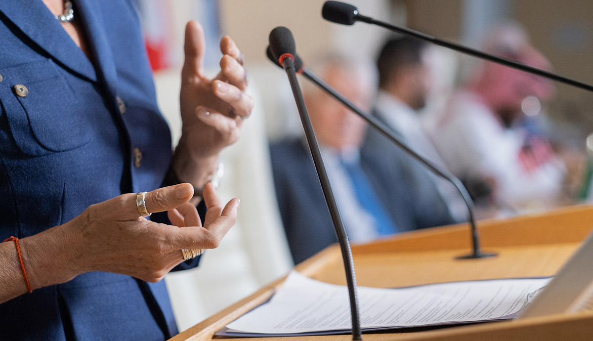 Speaker gesturing with hands at a wooden podium with multiple microphones during a presentation or meeting.