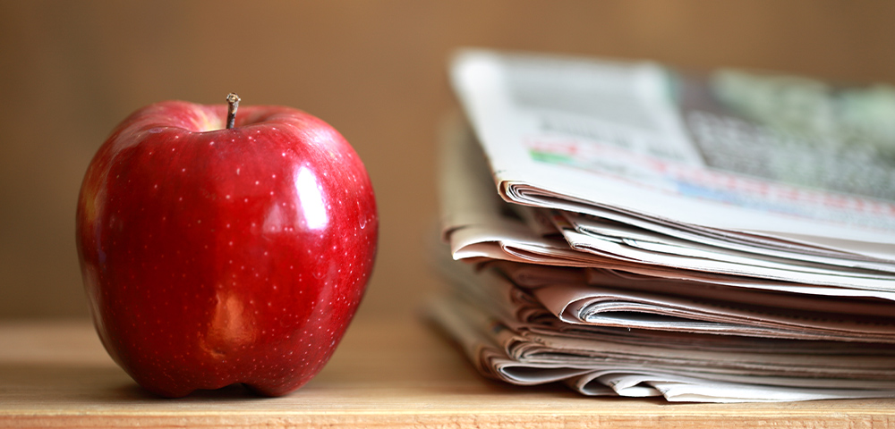 A red apple sits next to a stack of newspapers.