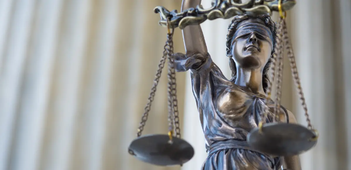 Landscape orientation digital stock cropped photograph close-up view of a dark bronze statue of Lady Justice, blindfolded and holding up a set of scales in her raised left hand; The background is a soft, light-colored curtain