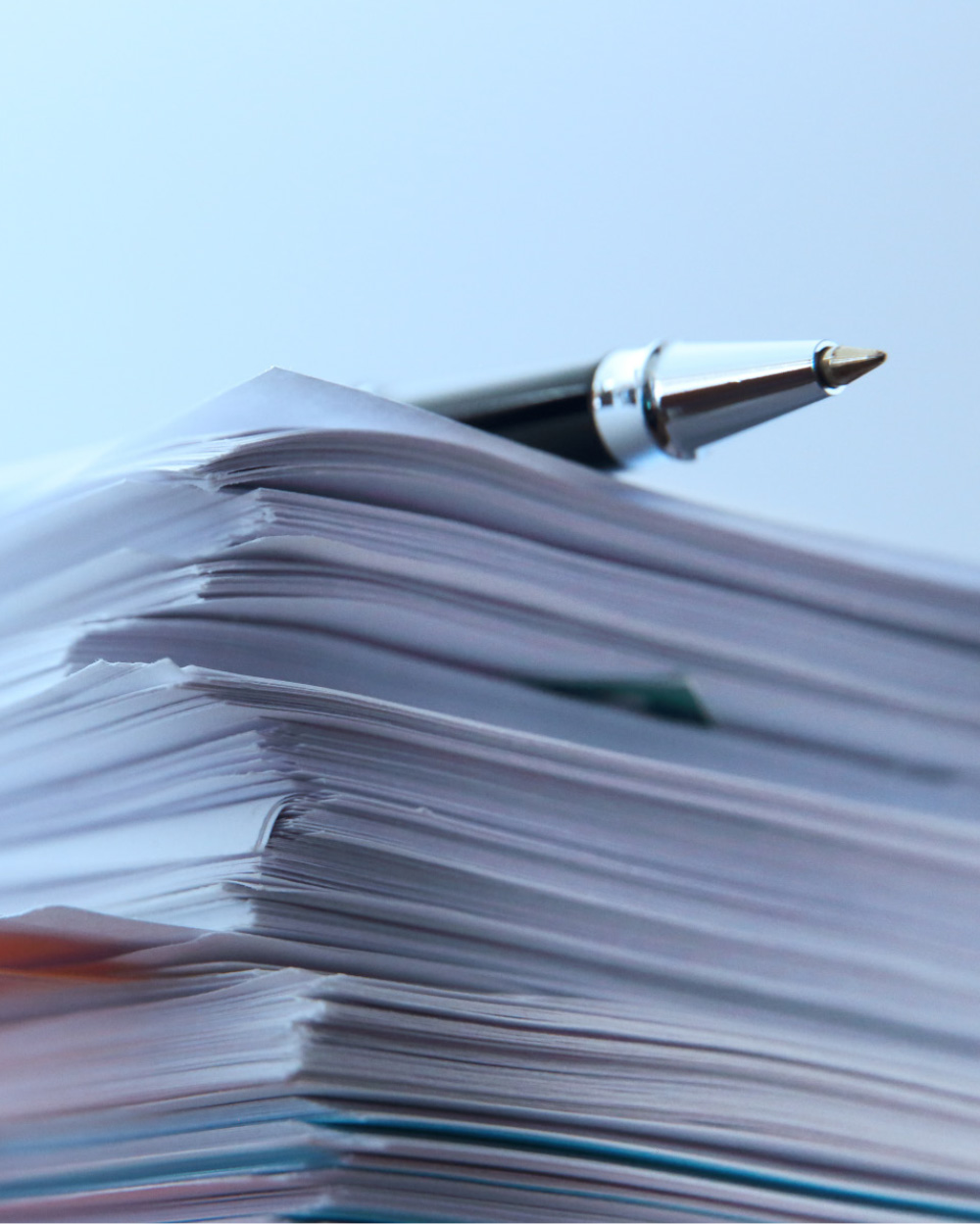 A high-angle, close-up shot of a thick stack of papers with a black and silver pen resting on top. The background is a soft, light blue.