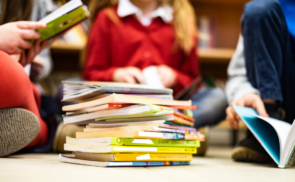 A low-angle close-up shot shows a stack of colorful children's books on a light-colored floor.