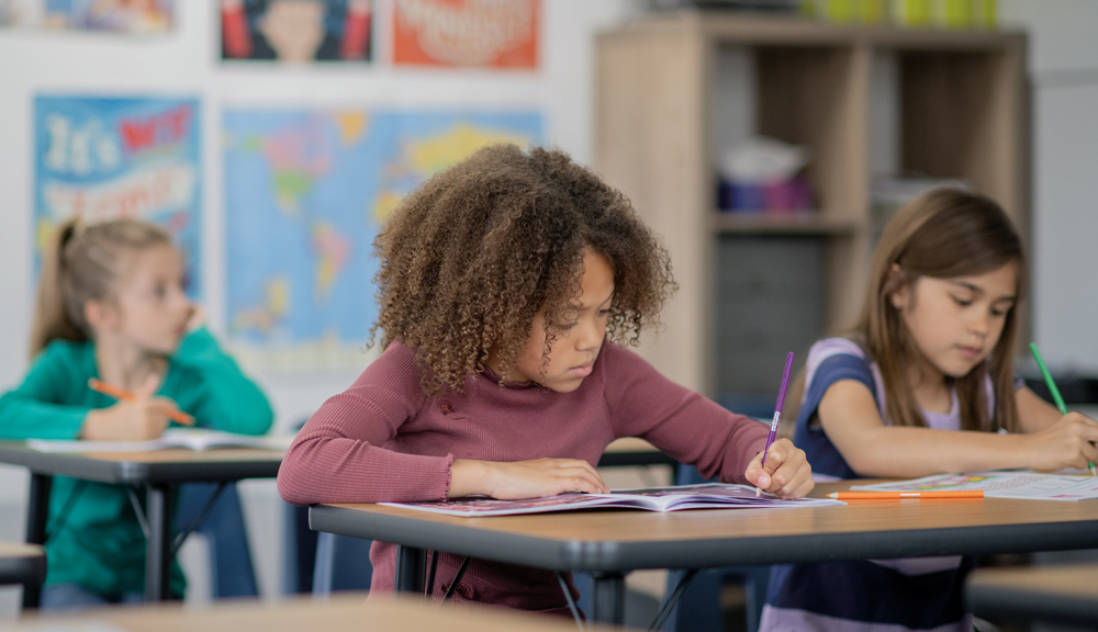Three children writing at desks in a classroom with educational posters in the background.