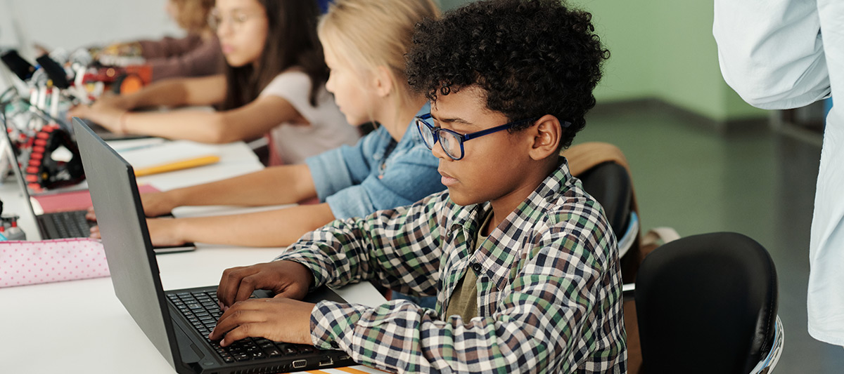 A focused young student, wearing glasses, works on a laptop in a classroom while other children sit at a long table in the background.