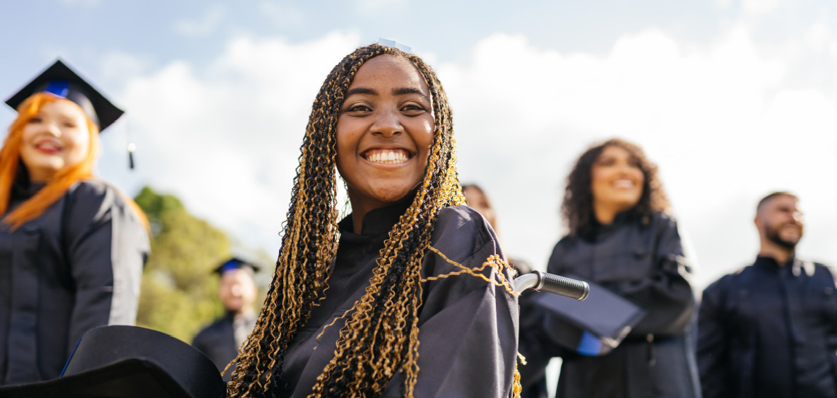 A close-up, eye-level shot of a smiling woman with long hair. She is wearing a dark graduation gown and cap.