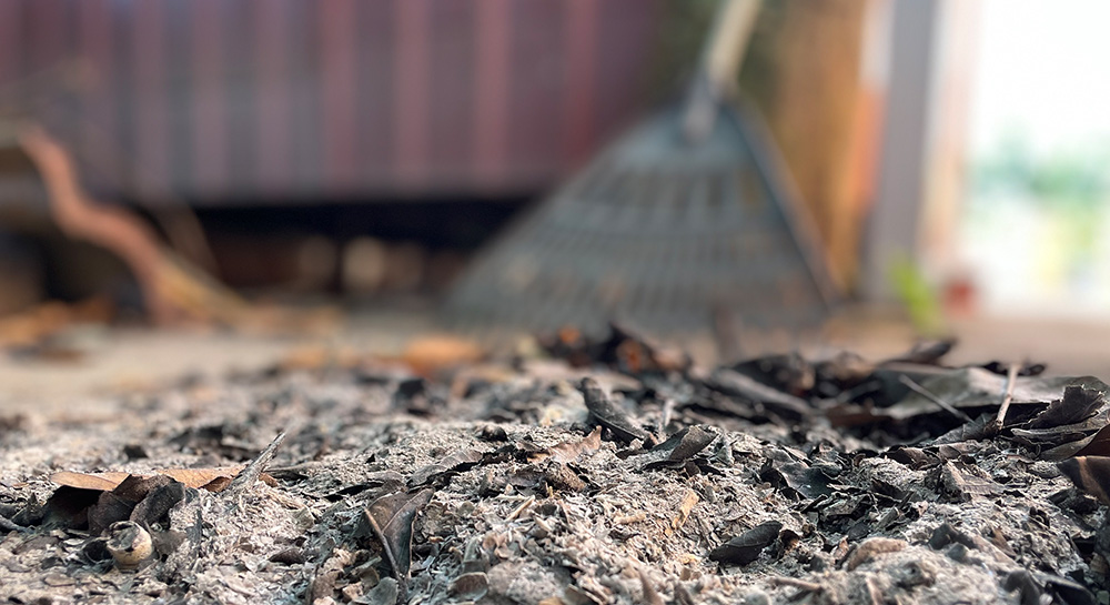 Close view of a pile of ash and debris, a rake leans against a wall, out of focus in the background.
