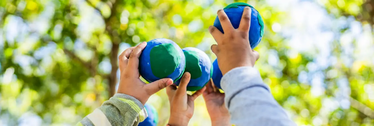 Landscape orientation digital stock cropped photograph close-up view of a group of children's hands reaching up into a leafy canopy of trees, each holding a small blue and green globe circular ball shape object, representing the Earth; The image is taken from a low angle, looking up towards the sky and trees