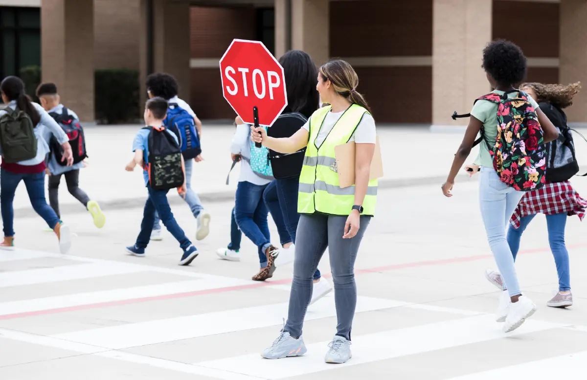 Landscape orientation digital stock photograph image display of a female crossing guard woman in a high-visibility vest and casual dressing attire holding a stop sign is standing in the middle of a crosswalk, observing and allowing a group of diverse elementary and middle school students in casual dressing attire with backpacks to safely cross the street in front of a school building