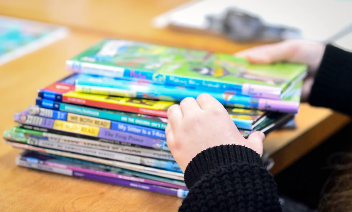 A student's hands are shown reaching for a stack of children's books on a wooden desk. The books have colorful covers with titles like "My Sister is a T-Rex" and "The Frog Prince." The student is wearing a black knit sweater.