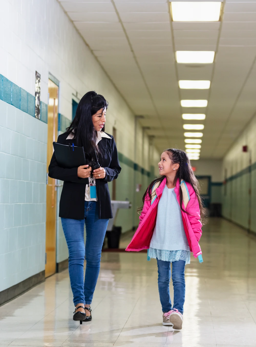 A teacher holding a clipboard and a young female student with a pink backpack are walking and talking in a school hallway. They are smiling at each other. The hallway has light blue walls and a white tiled floor and ceiling. The image is taken from a low angle.