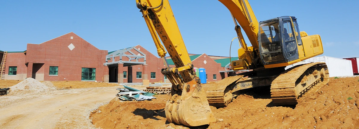 A large yellow excavator is parked on a dirt construction site, with its bucket resting on the ground in the foreground. In the background, a red brick school building is under construction, with scaffolding and other building materials scattered around. The sky is clear and blue.