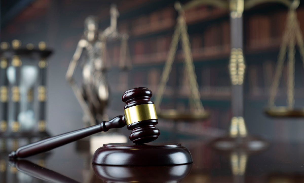 A low-angle, eye-level shot of a wooden gavel with a brass band, resting on a wooden sounding block. In the blurred background, a golden scale of justice and a statuette of Lady Justice are visible, positioned in front of a bookcase.