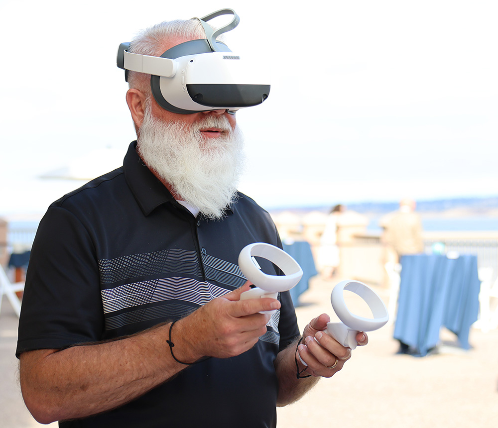 A man with a white beard wears a virtual reality headset and holds two controllers.