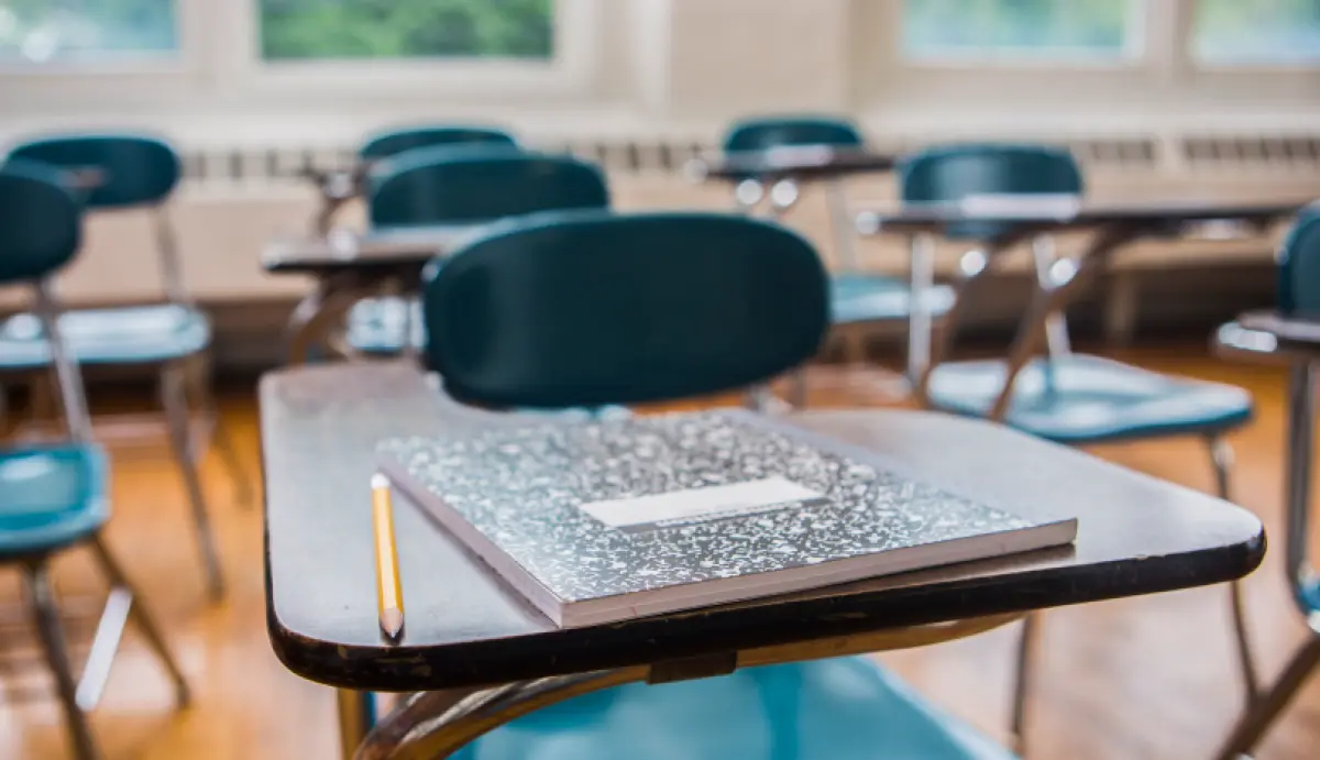 Landscape orientation digital stock photograph image display of a low, close-up shot of a vintage wooden school desk with an attached teal colored chair; A composition notebook with a speckled black and white cover and a wooden pencil are on the desk; The background is a blurred view of a classroom with rows of similar desks attached with teal colored chairs and large windows are situated behind everything