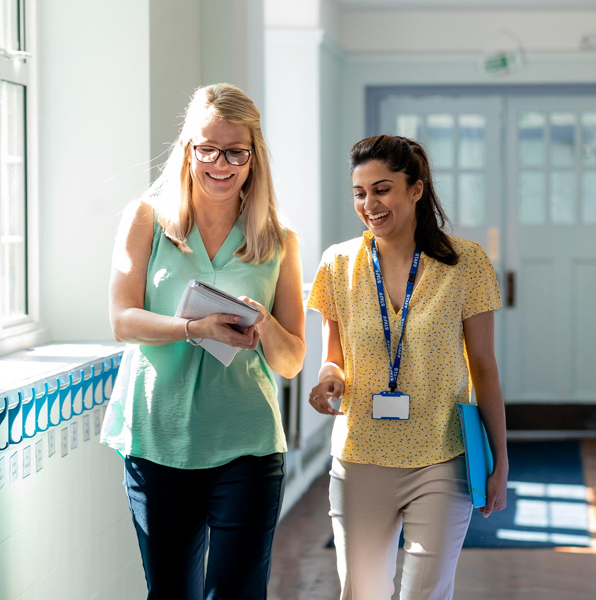 Two smiling female teachers or school administrators walking together and collaborating in a bright school hallway