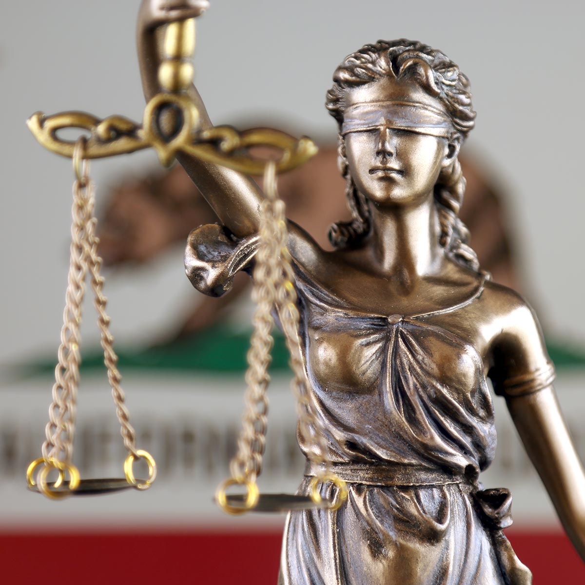 Bronze statue of Lady Justice holding scales, symbolizing law and justice, with the California State Flag blurred in the background