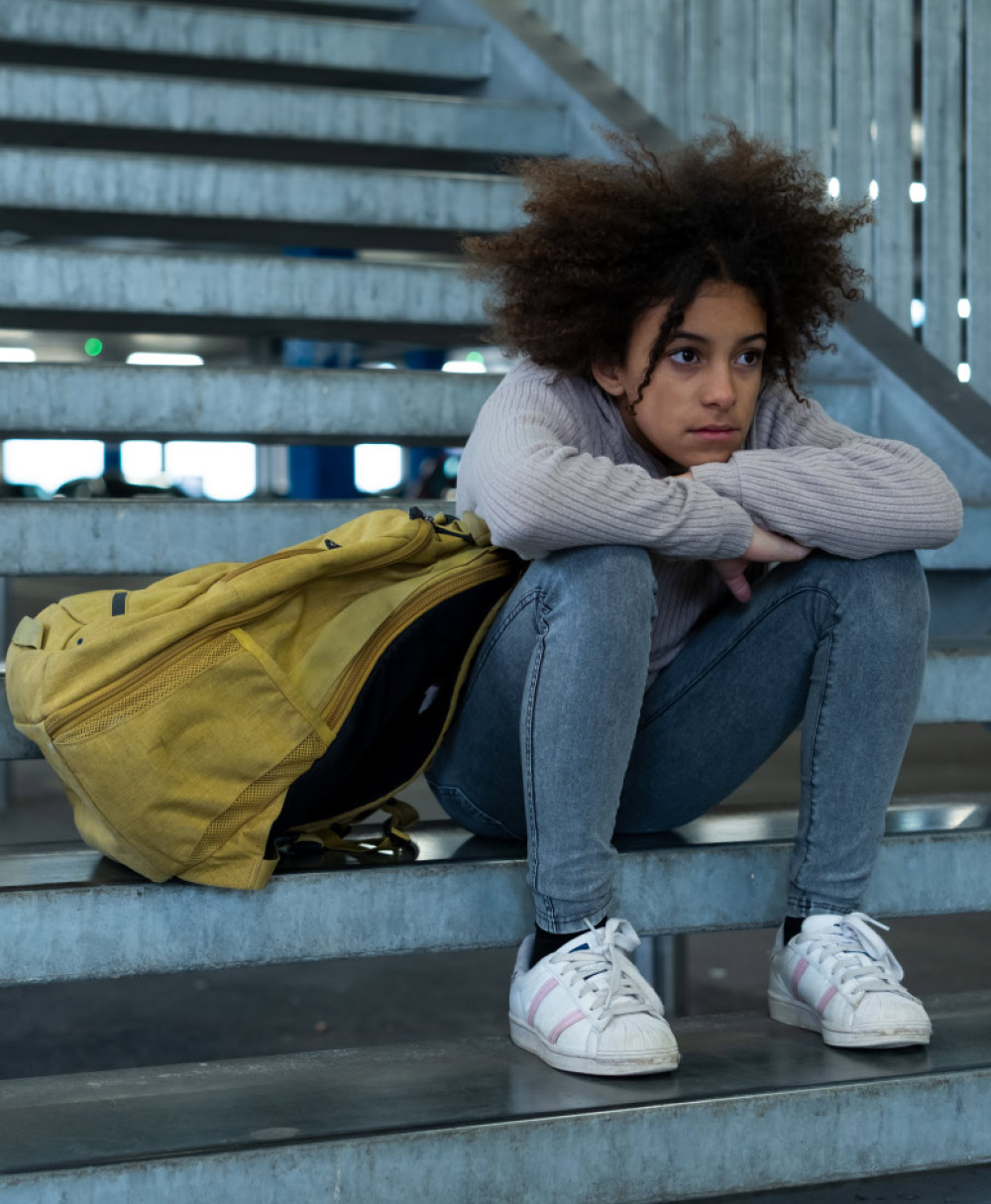 A young girl with curly hair is sitting alone on metal stairs with a sad or worried expression. She is leaning over her knees next to her yellow backpack. The setting looks like an outdoor stairwell.