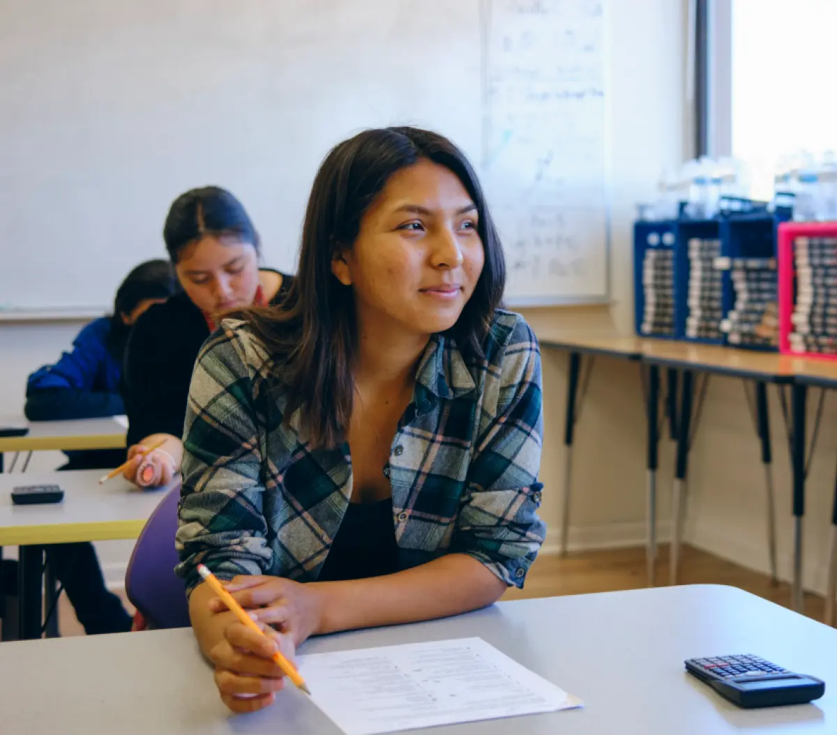 A close-up photograph cropped portrait orientation view of a female student seated at a desk in a classroom looking toward the bright window on the right; She is wearing a multi-colored (shades of teal blue, white, and faded pink) plaid open button-up dress shirt and holding a pencil over a paper on her desk; A calculator is nearby; Other students are seated behind her, and in the background there is a whiteboard and a table holding stacks of bins and boxes