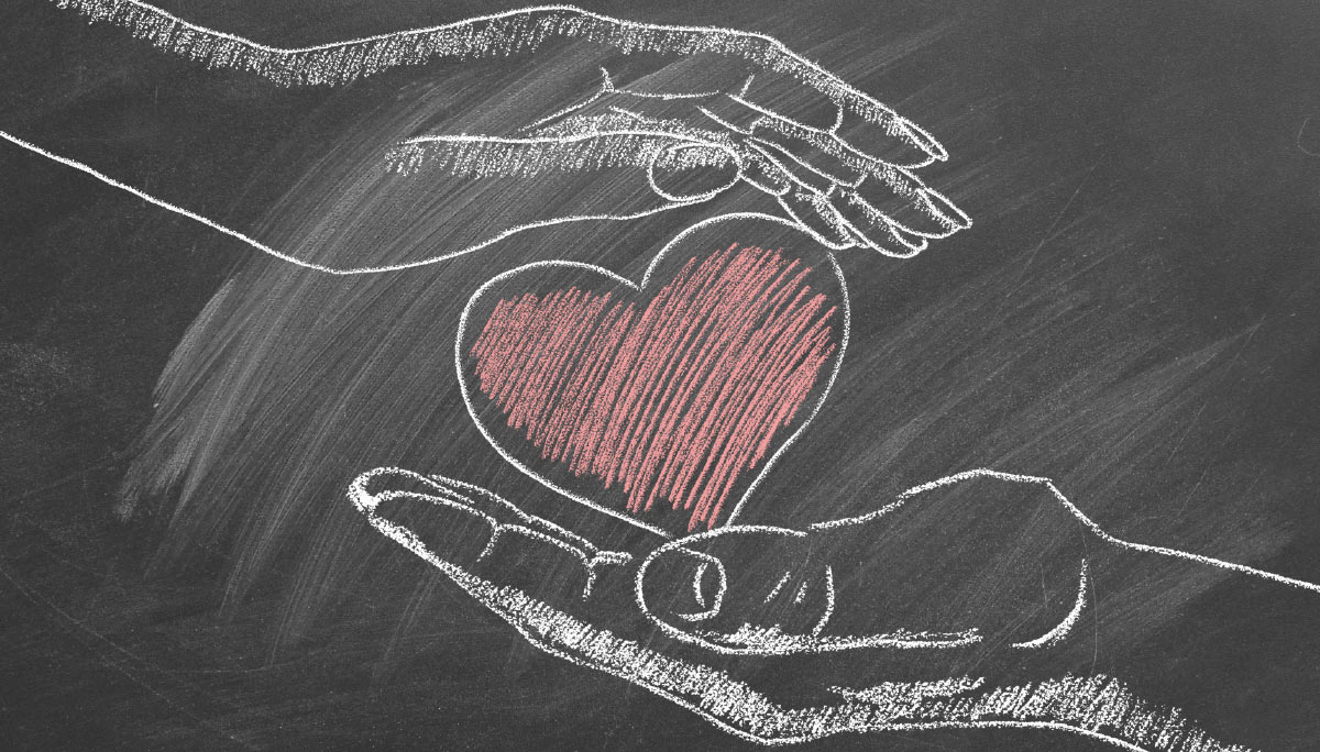 Chalk drawing on a blackboard showing two hands gently holding a large, scribbled red heart, symbolizing care or compassion.