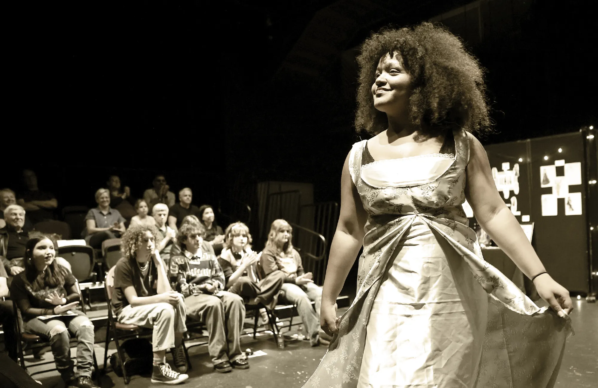 A young woman in a light-colored dress smiling brightly while performing on a stage for an audience.