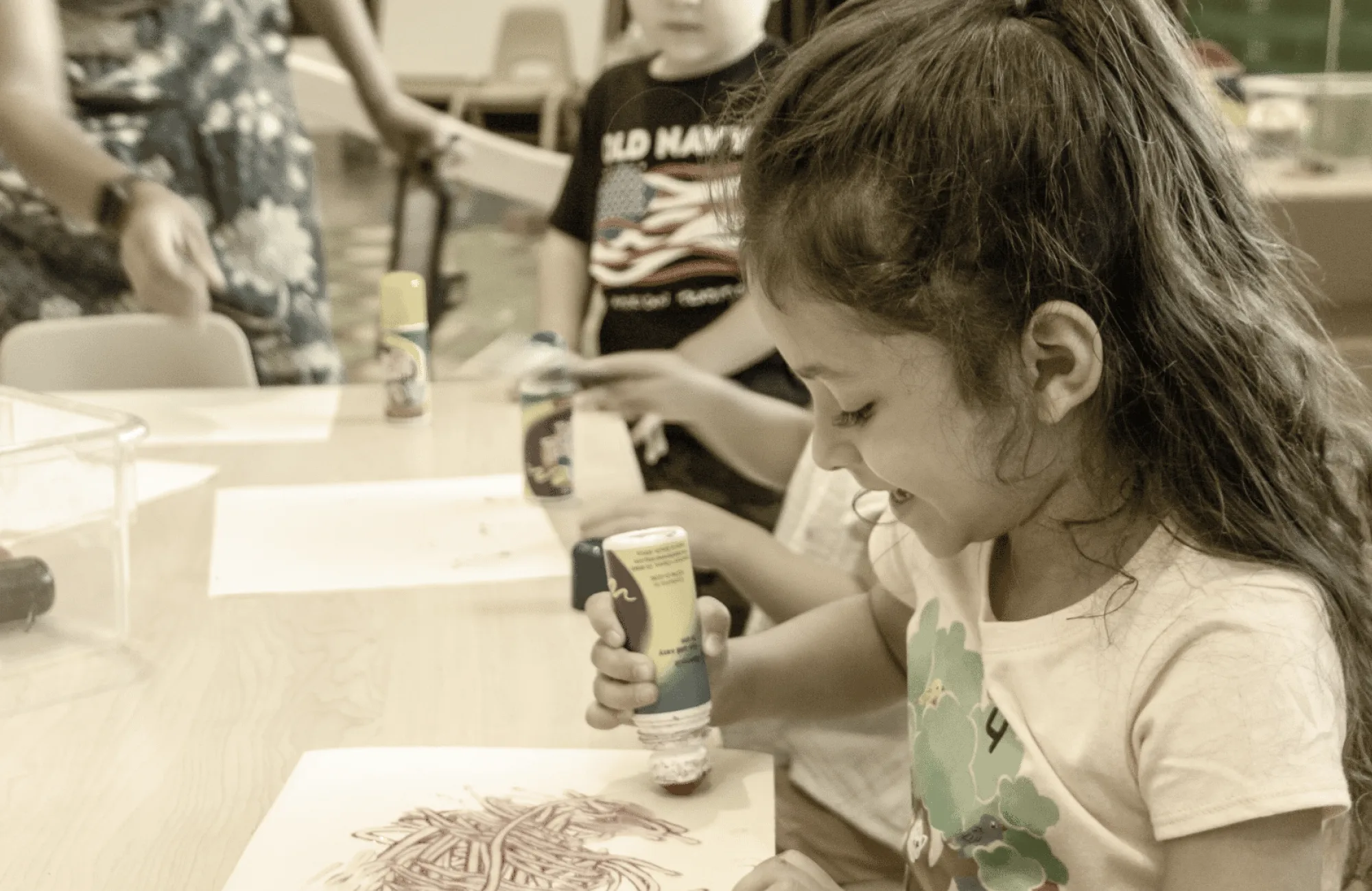 A young girl concentrating and smiling while using a glue stick for an art activity at a table with other children.