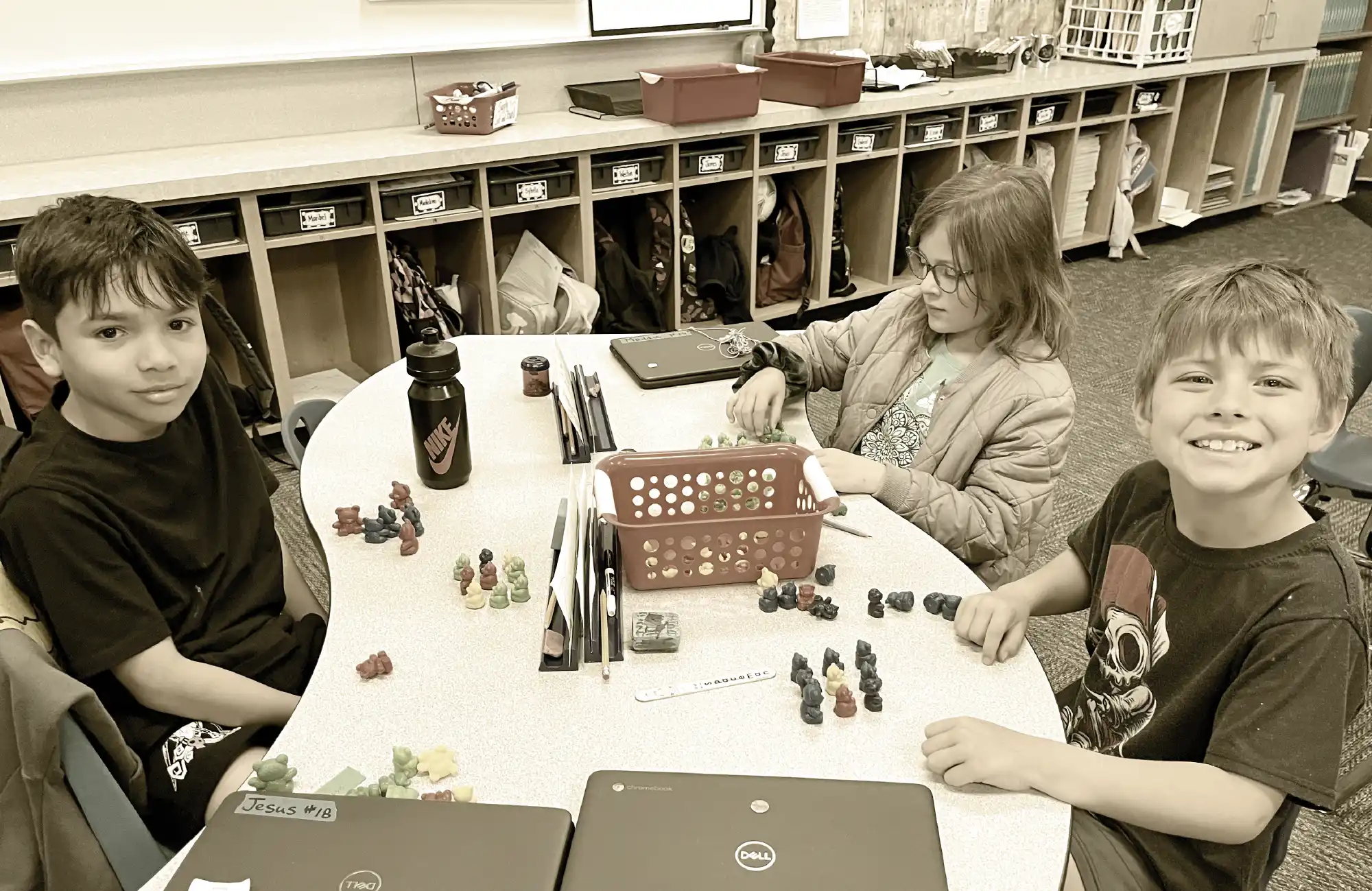 Three elementary students smiling while working with small figurines and Chromebooks at a table in a classroom.