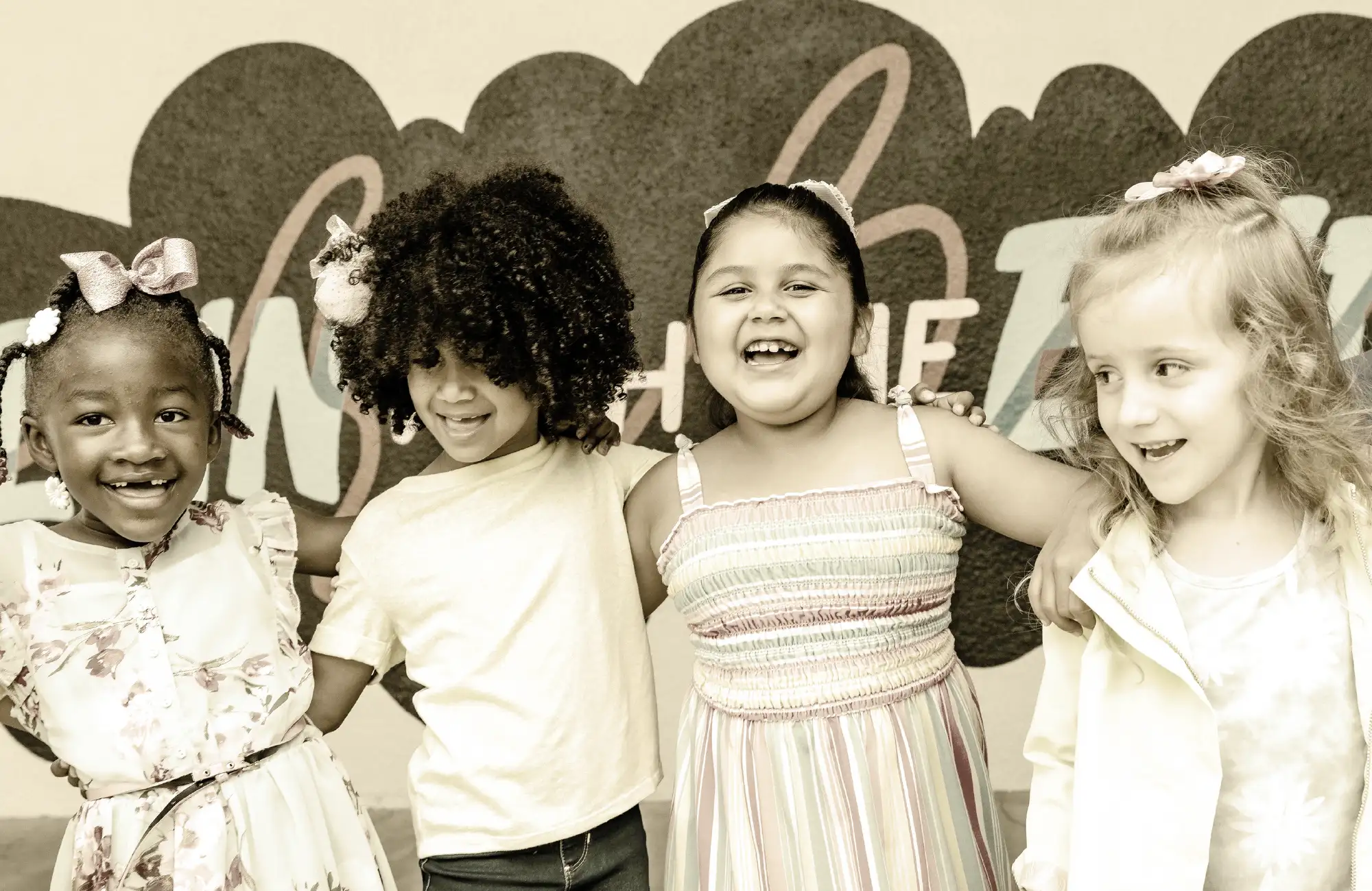 Four young girls standing side-by-side with their arms around each other, laughing happily.