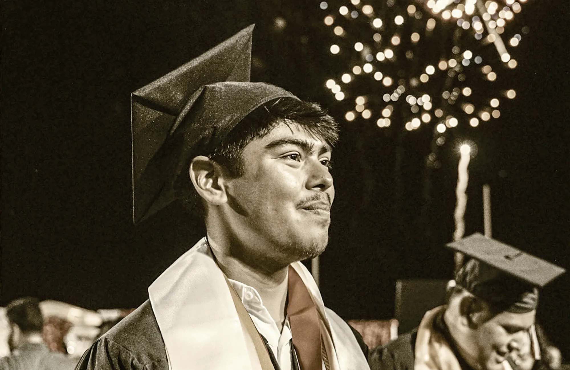 A male high school graduate smiling in his cap and gown at night, with fireworks exploding in the blurred background.