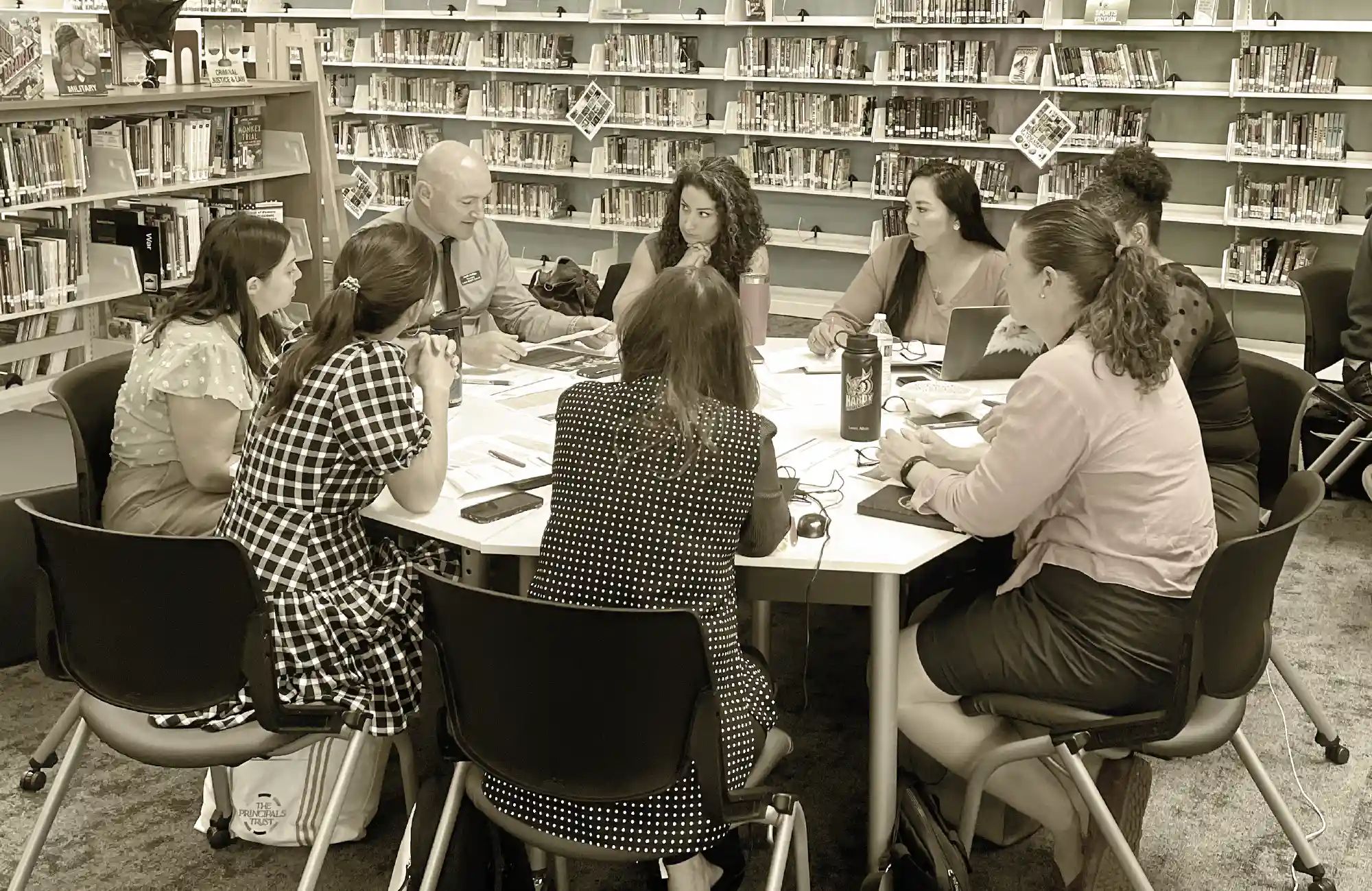 A group of eight educators and one male administrator seated around a table for a meeting in a library.