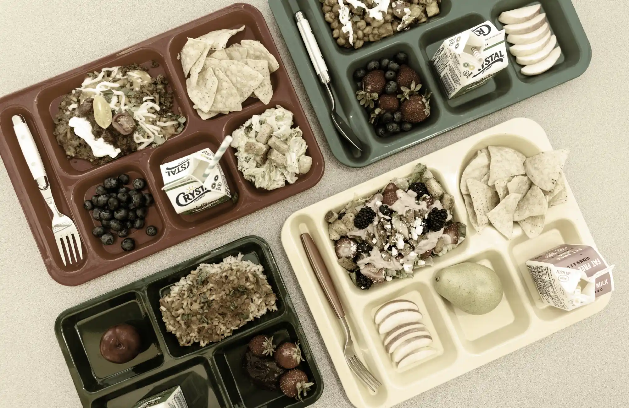 Overhead view of four school lunch trays containing various meals, including salads, fruit, rice, chips, and milk cartons.