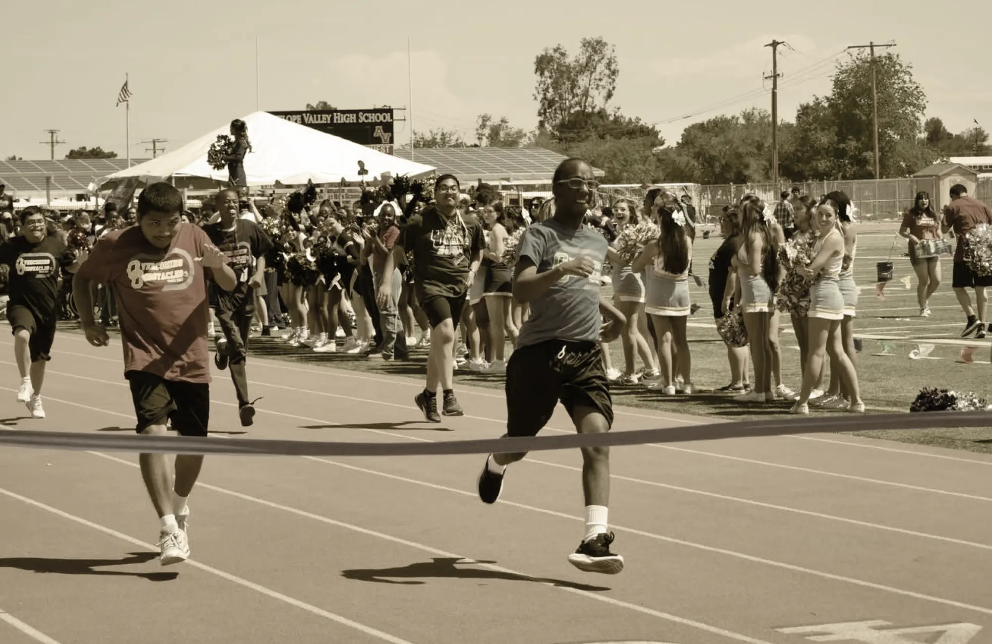 People running toward the finish line on a track, with cheerleaders and spectators lining the field.