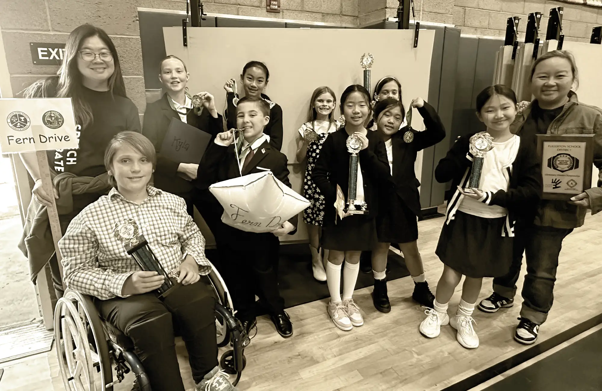 A group of elementary students and two adults, proudly holding trophies, medals, and awards indoors.