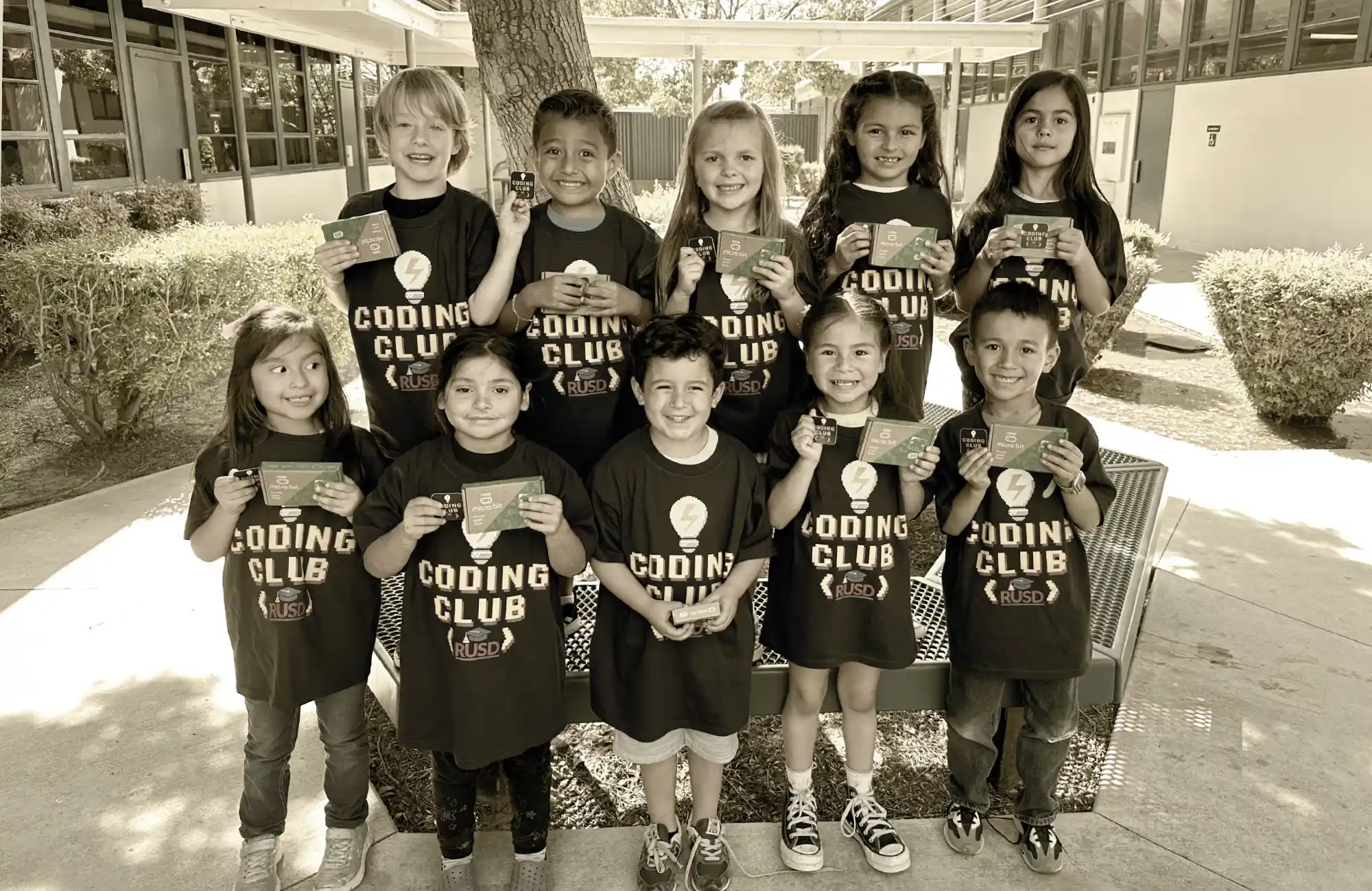 A group of ten smiling elementary school students wearing "Coding Club RUSD" t-shirts and holding cards outdoors.