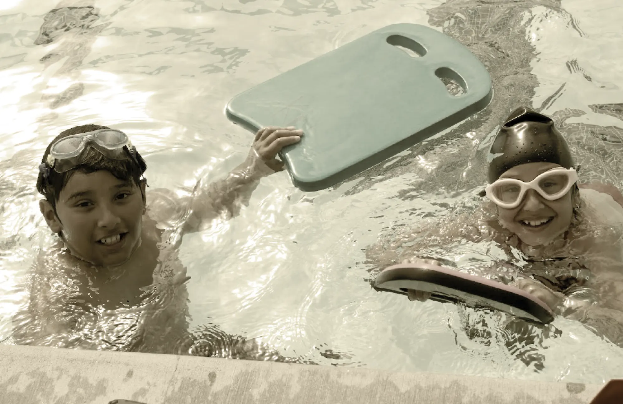 Two smiling children wearing goggles and swim caps, holding kickboards in a swimming pool.