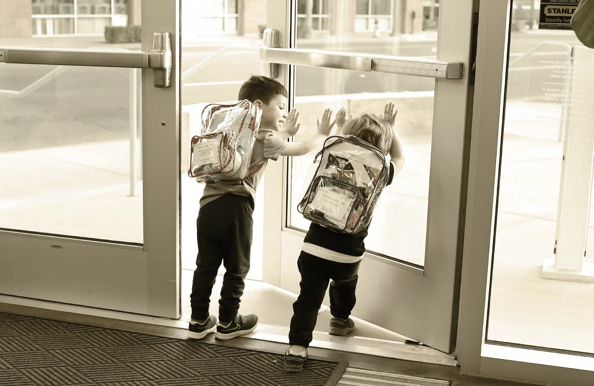 Two young boys with backpacks, pressing their hands against a glass school door as they look outside.