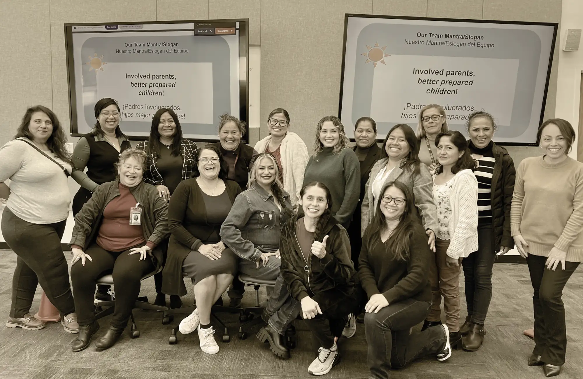 A group photo of 15 smiling women in front of screens displaying the mantra: "Involved parents, better prepared children!"