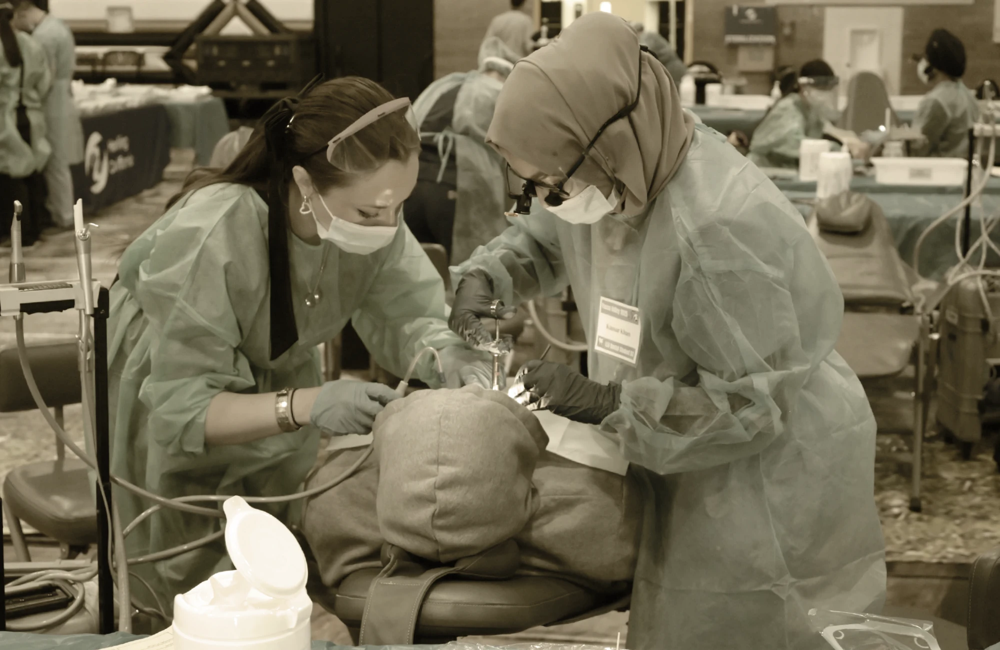 Two female dentists/hygienists in gowns, masks, and gloves performing dental work on a patient lying down.