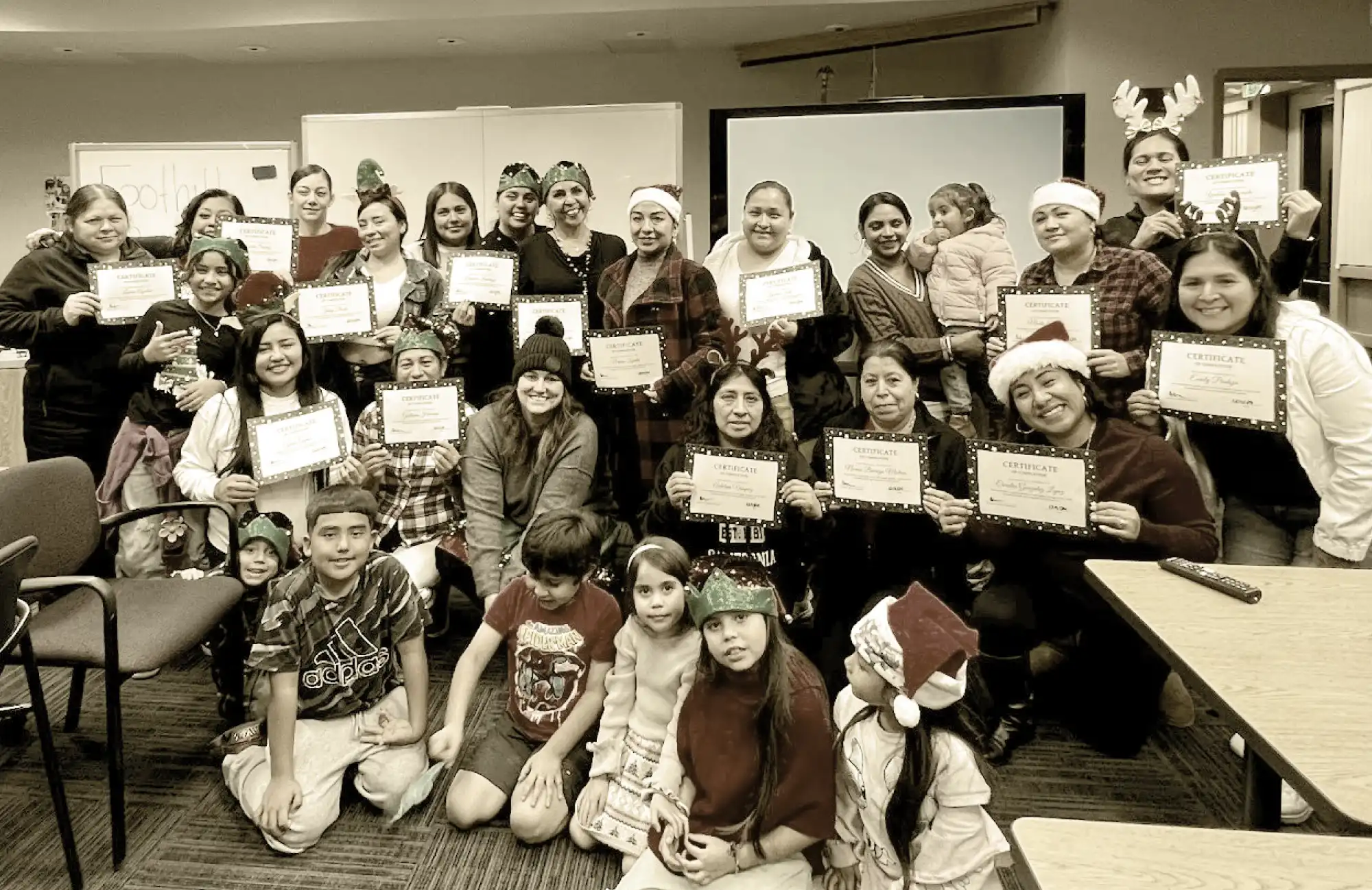 A group photo of women and children wearing holiday hats and happily holding up "Certificate" awards indoors.