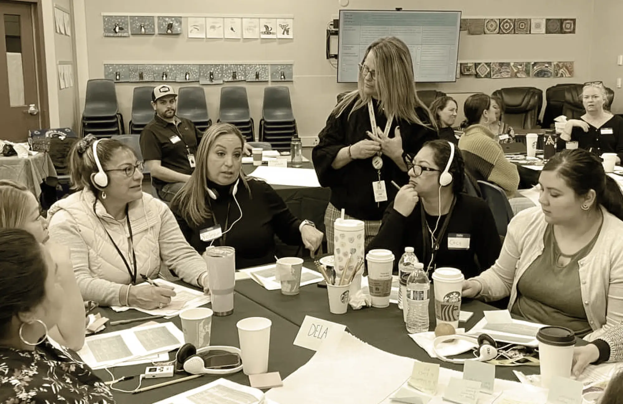 A group of adults in a training session, seated at tables and wearing headphones for simultaneous translation.