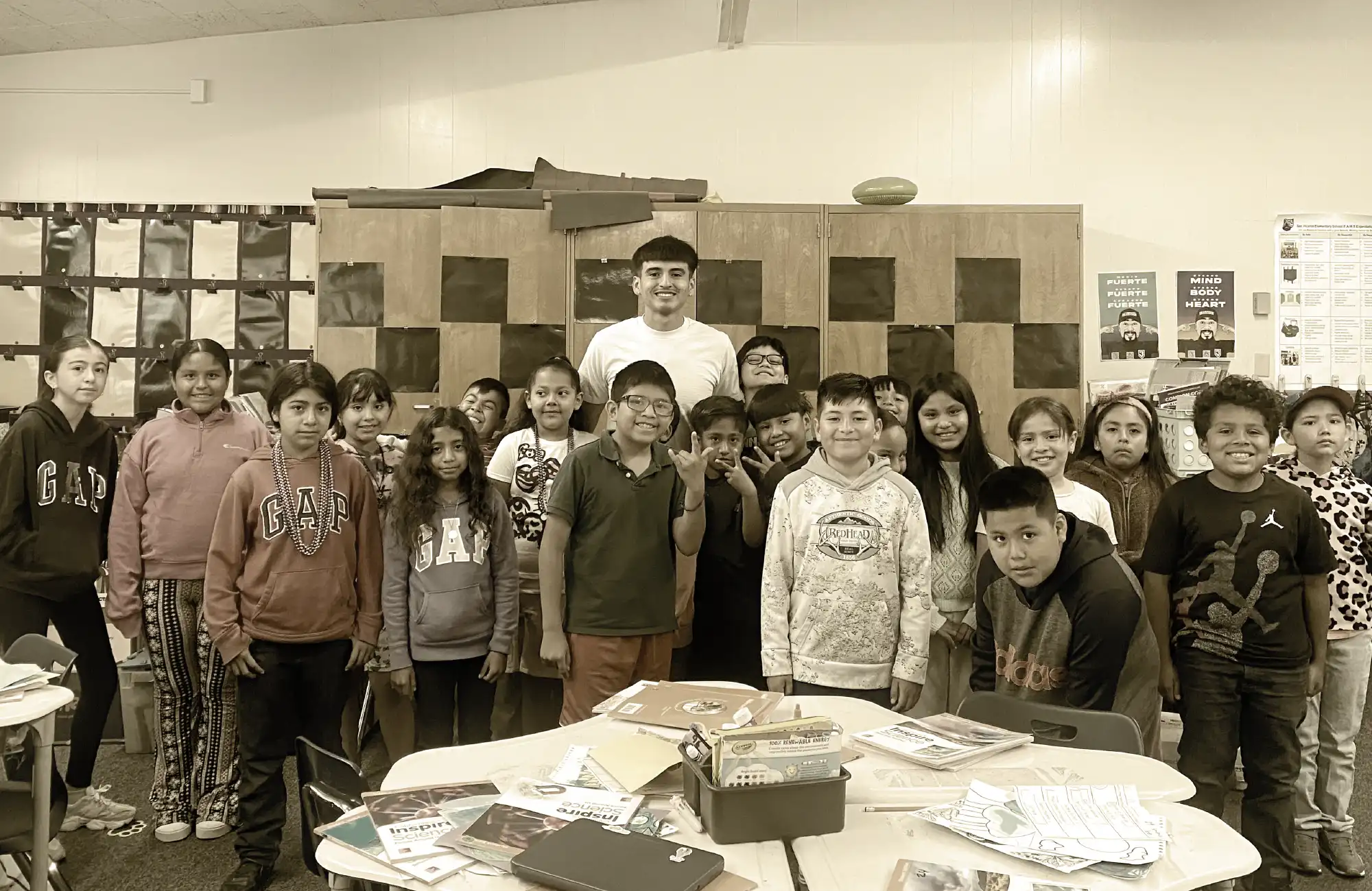 A male high school student standing with a group of over a dozen elementary school children in a classroom.