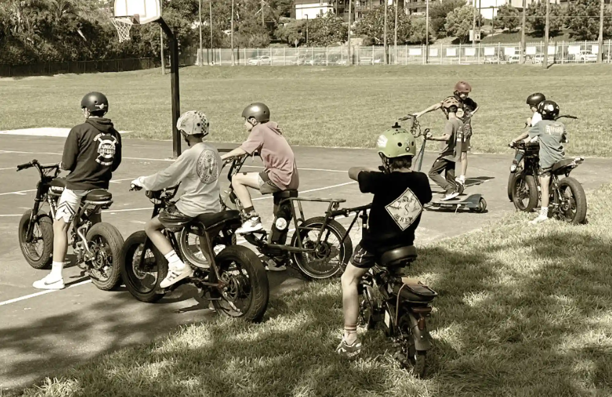 Children wearing helmets and riding electric bicycles or mini-bikes on a sunny playground/field.