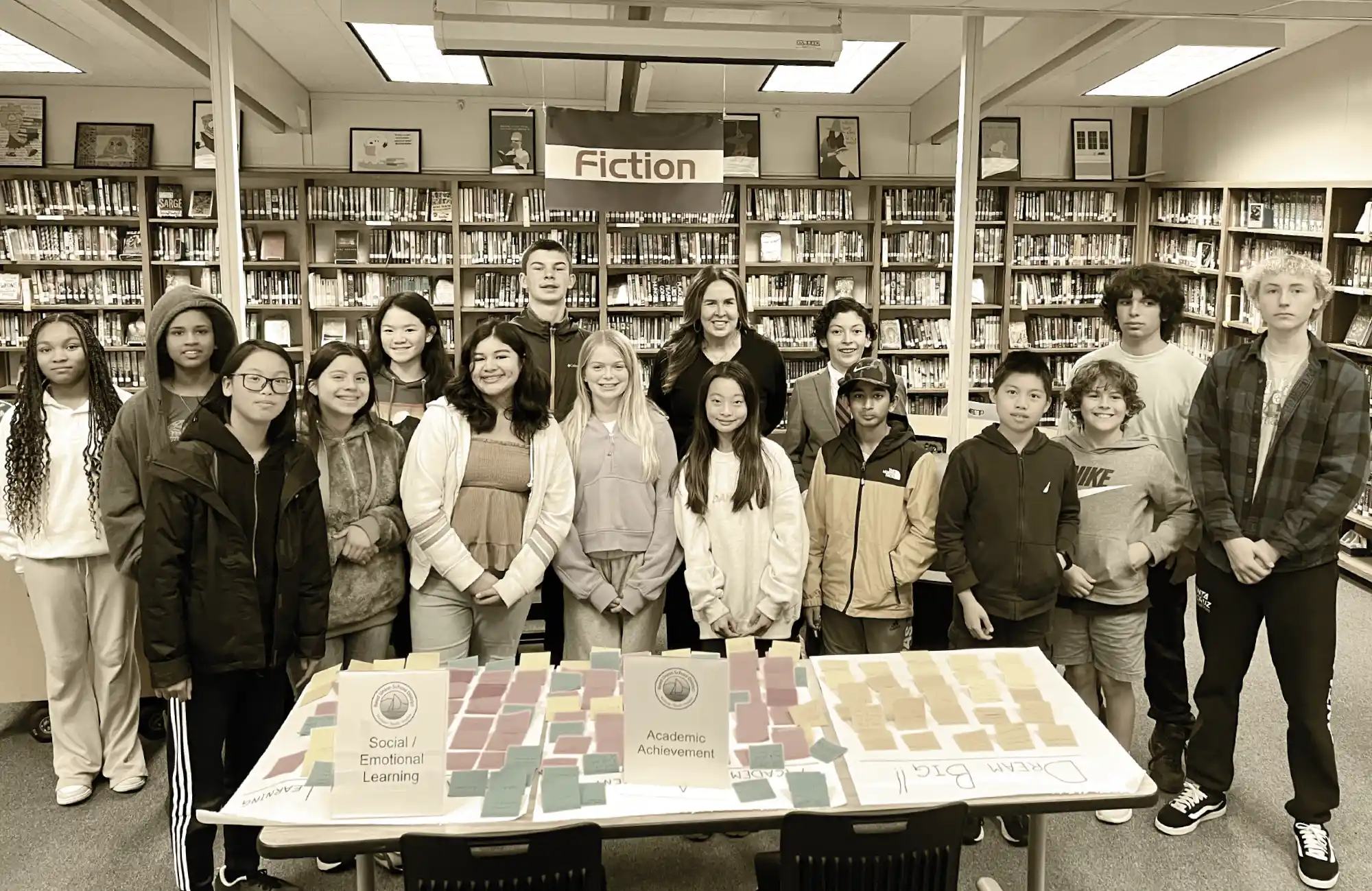 A group photo of a teacher and students standing in a library behind a table of awards/cards for "Academic Achievement" and "Social/Emotional Learning."