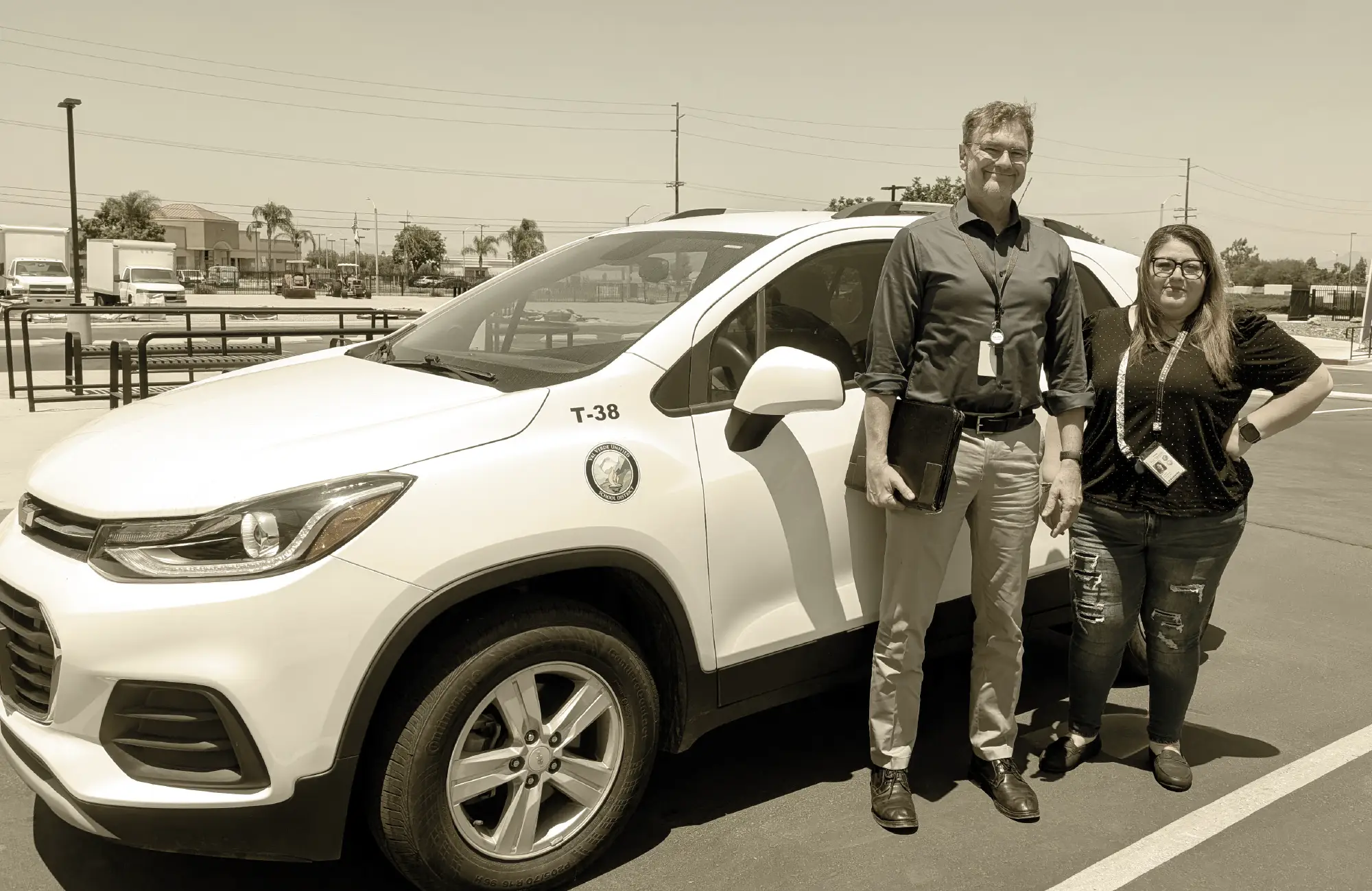 A man and a woman standing next to a white school police or security vehicle labeled T-38 in a sunny parking lot.