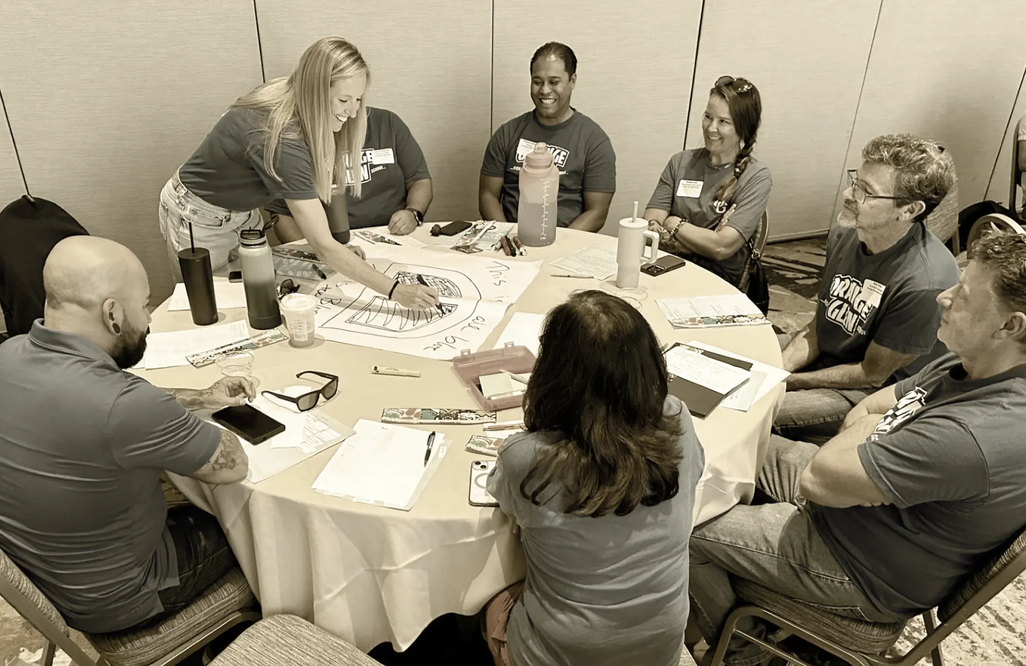 Seven adults participating in a professional learning activity around a circular table, drawing on a large paper.