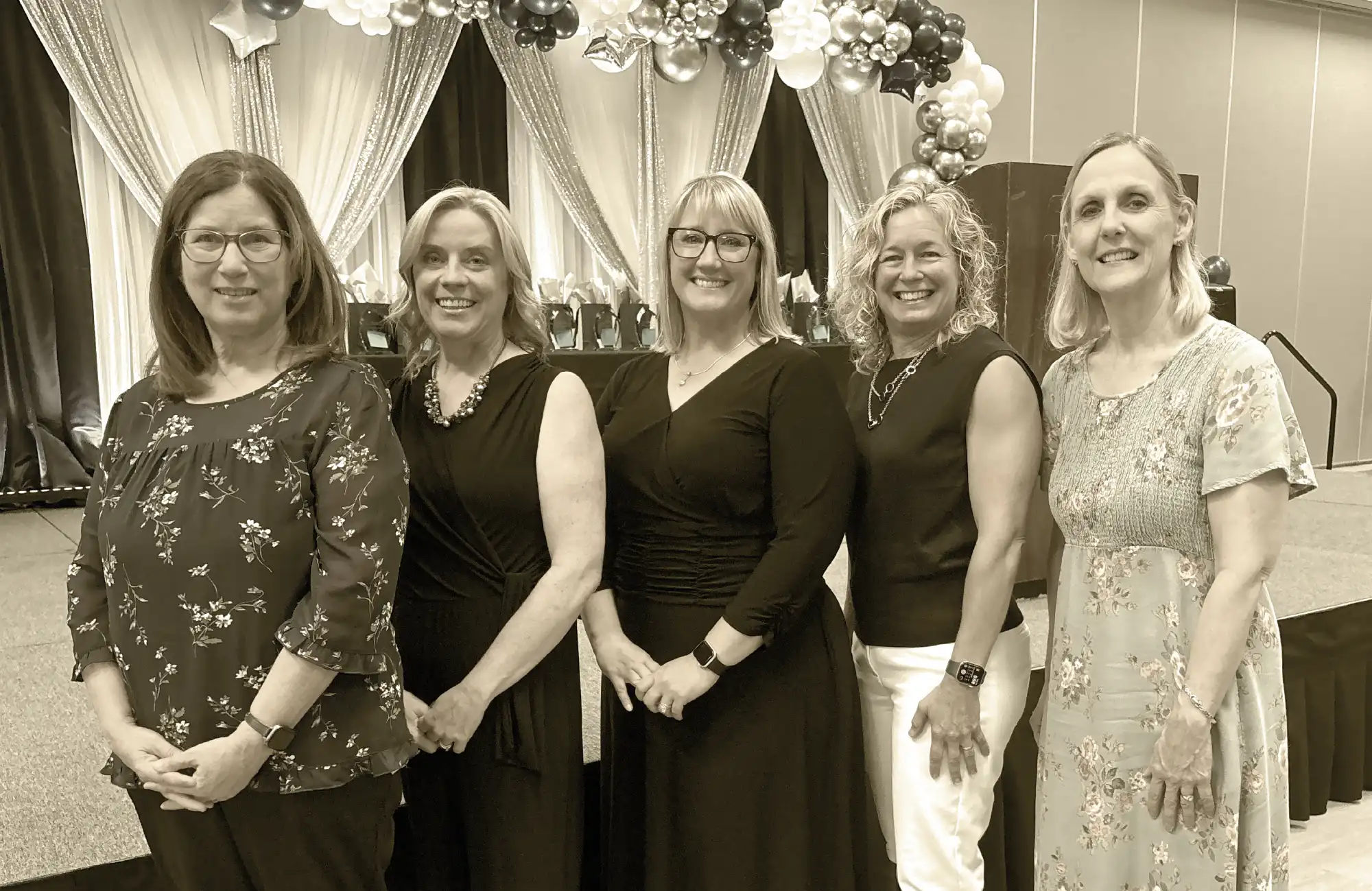 A group portrait of five smiling women dressed professionally, standing on a stage in front of a backdrop with balloons.