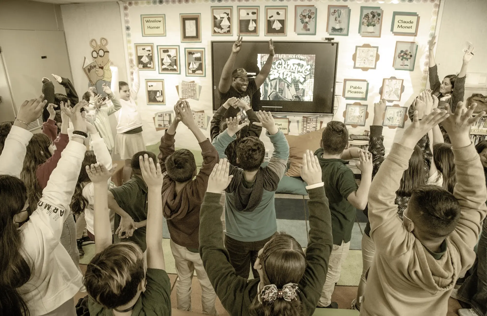 Students reaching their hands up toward the ceiling in an active, collaborative classroom setting.