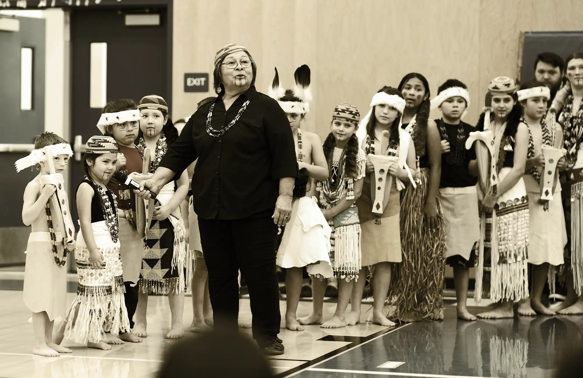 Performance featuring an elder leading a line of children wearing white fringe and feather headbands.