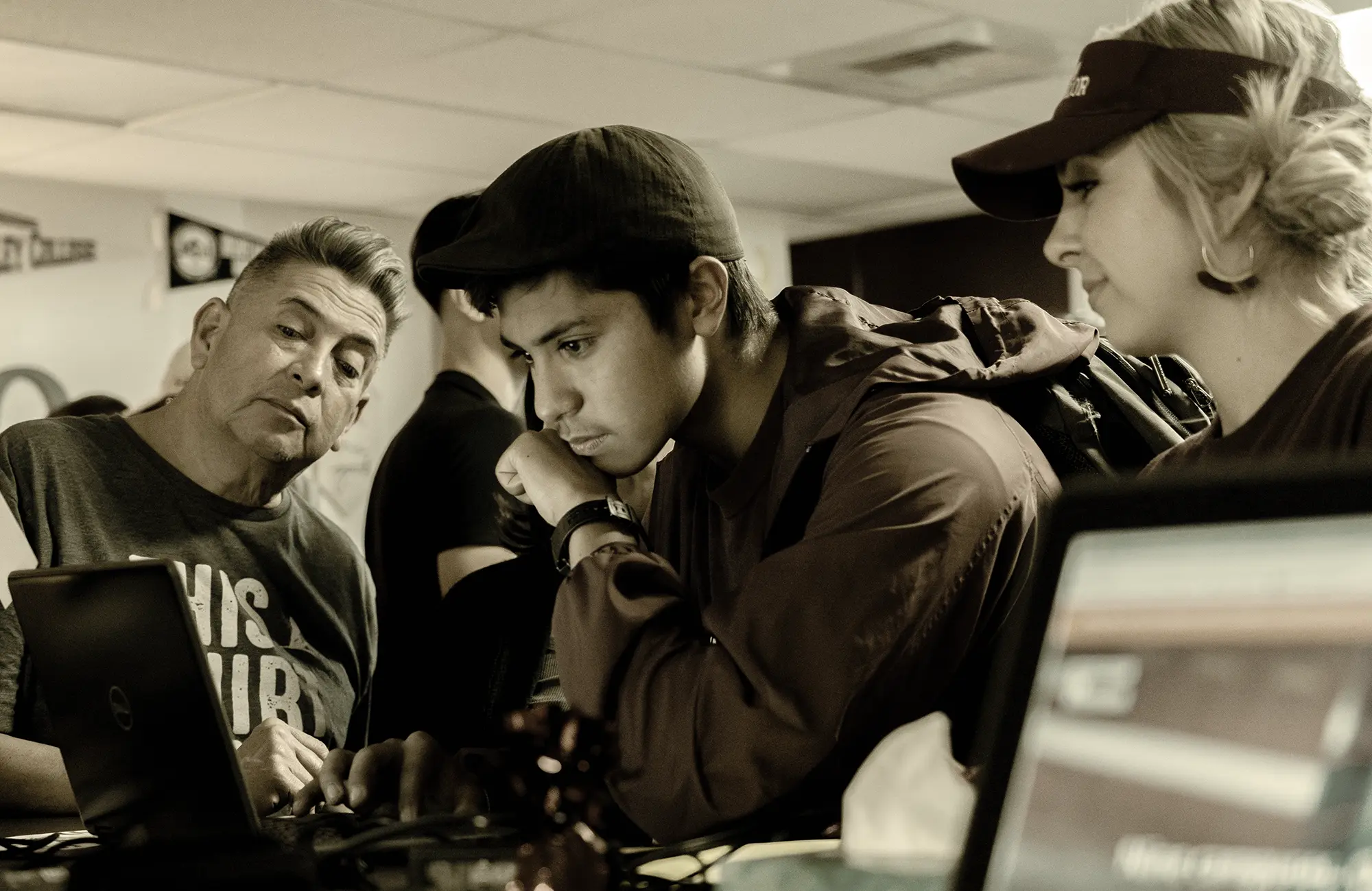 High school student receiving guidance from two adult mentors while working on a laptop in a computer lab.